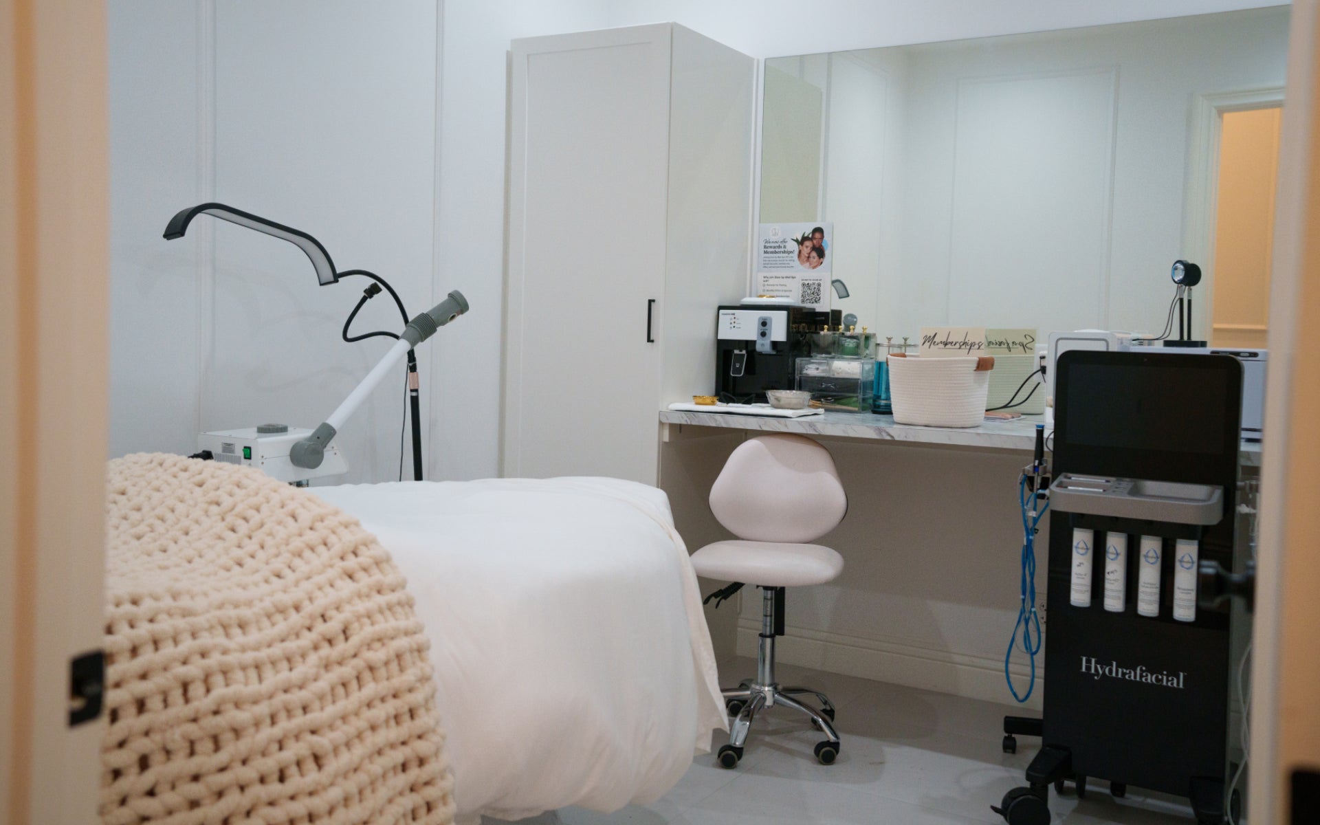 A brightly lit, clean, and modern med spa treatment room, featuring a white treatment bed with a textured blanket, a supply counter, and a black cart with a HydraFacial machine.