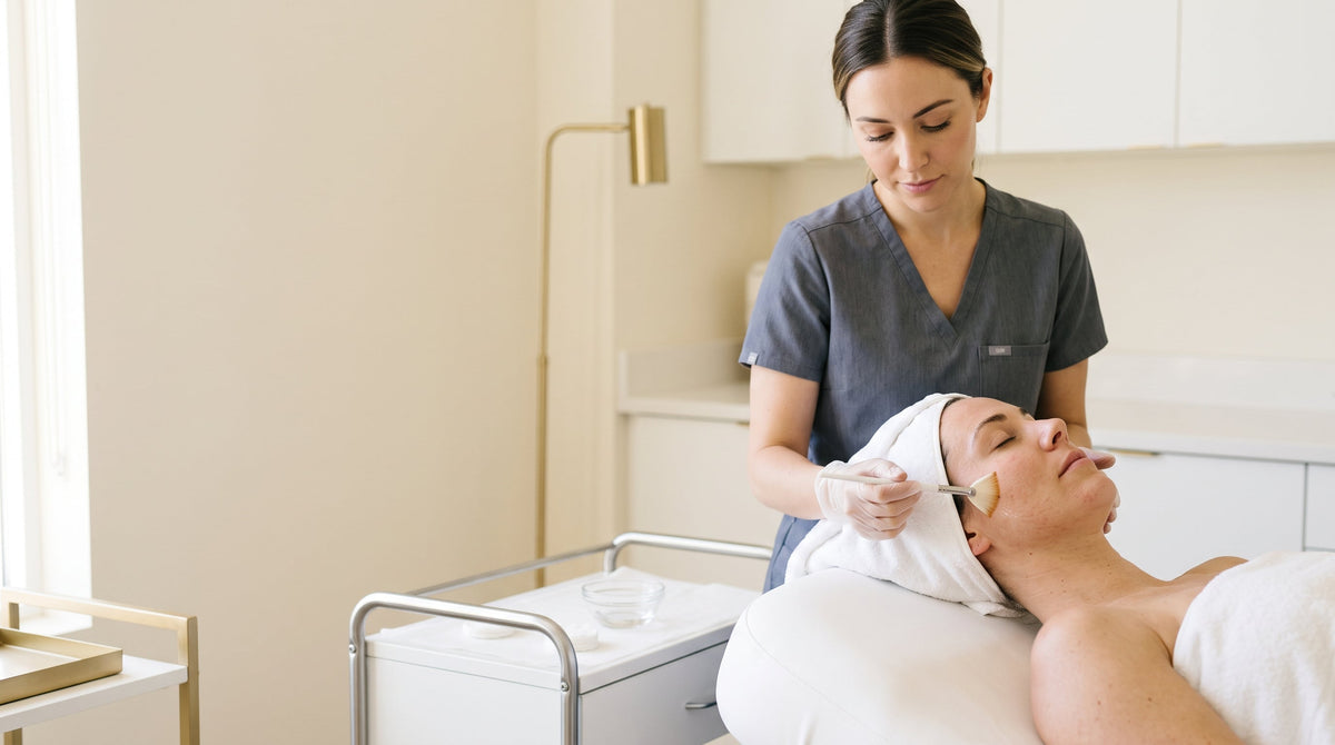 Provider in grey scrubs brushing a clear peel solution onto a relaxed client's cheek in a bright room